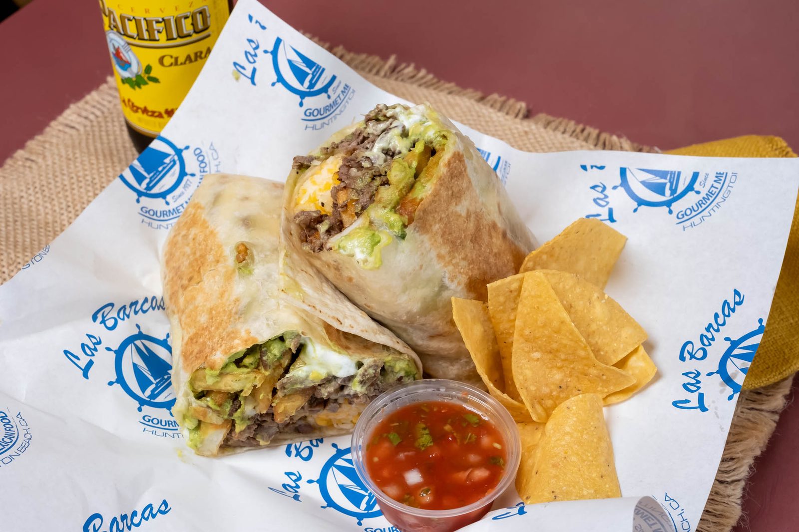 A burrito cut open to show its fillings, served with chips and salsa on Las Barcas' branded paper.