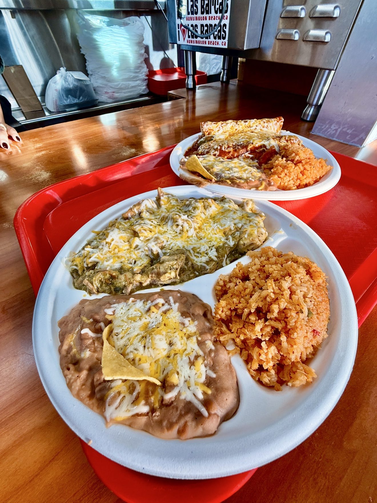 Two combination plates on a red tray at the Las Barcas counter: chile verde in green sauce and the enchilada plate in red sauce, each served with rice and refried beans.